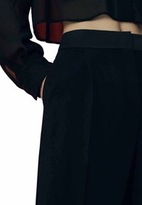 Pantalons noirs à jambes larges avec une texture lisse et une taille sur mesure. Dotés d'une poche latérale, ils sont associés à un haut semi-transparent noir.