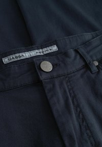 Detalj tamno mornarsko plavih tkanih hlača koji prikazuje pojas, gumb s nazivom marke "Gabba" i unutarnju etiketu s markom.