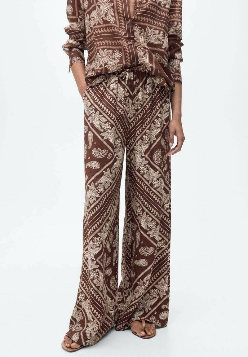 Personne portant un pantalon large marron et une chemise assortie avec des motifs paisley beiges, les mains dans les poches et des sandales aux pieds.