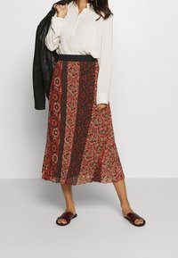 Midi-jurk met bloemenpatroon in rood en zwart, met een vloeiende uitstraling en een brede zwarte tailleband. Gemaakt in combinatie met een witte blouse en zwarte sandalen.