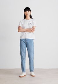 T-shirt bianca in cotone con tasca, jeans in denim blu e sneakers bianche con suola marrone. Modello in piedi con le braccia incrociate su uno sfondo chiaro.