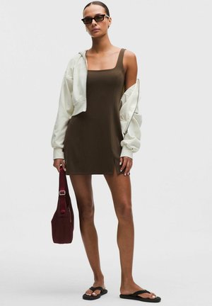 Mujer con vestido marrón sin mangas, chaqueta corta blanca caída de un hombro, gafas de sol negras, sandalias negras y sujetando un bolso granate.