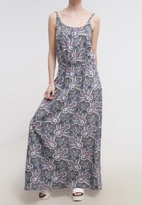 Kobieta w bezrękawnej sukience maxi w niebieski, biały i różowy wzór kwiatowy paisley oraz białych sandałach na platformie.