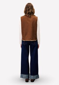 Person mit welligem Haar, der eine braune ärmellose Weste über einem cremefarbenen Langarmshirt, weit geschnittener, dunkelblauer, umgeschlagener Jeans und braunen Schuhen trägt und steht.