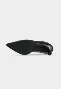 Chaussure à talon haut noire à bout pointu, posée sur le côté, montrant une semelle lisse et un talon fin sur un fond uni.