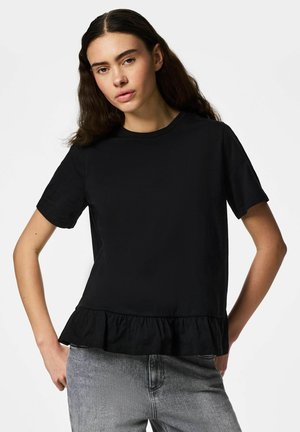 Jauna sieviete ar gariem tumšiem matiem valkā melnu īss piedurkņu topu ar rievotu apakšmalu un gaiši pelēkas džinsa bikses uz balta fona.