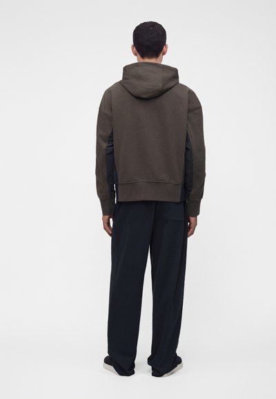 Homme debout, de dos, portant un sweat à capuche vert olive foncé et un pantalon noir ample avec des chaussures noires sur un fond clair uni.