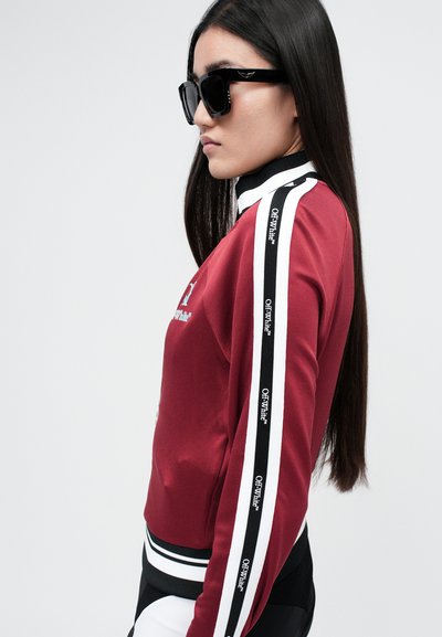 Giacca sportiva rossa con dettagli a righe nere e bianche, collo alto, logo frontale e una texture liscia. Il modello indossa occhiali da sole neri.