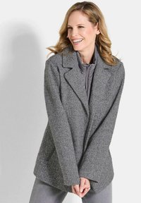Frau lächelnd, trägt einen grauen Fischgrätmantel über einer gesteppte Zip-Jacke, steht vor einem schlichten hellen Hintergrund.