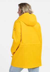 Gul vattentät jacka med huva, med cinchad midja och långa ärmar. Slät textur, minimal hårdvara och enkel design.