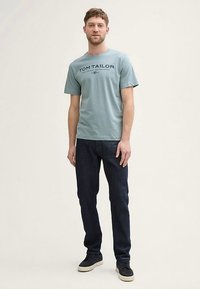 Vaaleansininen puuvillainen t-paita, jossa on tummilla kirjaimilla painettu 'TOM TAILOR', yhdistetty tummiin farkkuihin ja mustiin lenkkareihin. Yksinkertainen rento ilme.