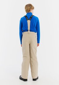 Beige sneeuwbroeken met verstelbare zwarte bretels, rechte snit en een gladde textuur, gedragen over een blauwe langemouwen top. Zwarte laarzen zichtbaar.