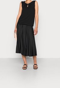 Top nero senza maniche con dettaglio a chiave, abbinato a una gonna midi plissettata nera e sandali piatti neri, tenuto con una clutch rosa.