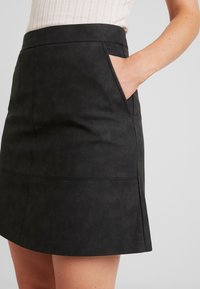Gonna nera in ecopelle con vita aderente, tasca laterale e dettagli di cucitura, caratterizzata da una texture liscia e da una forma leggermente svasata.