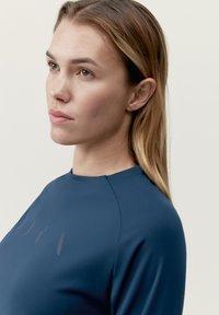 Marinblå plagg med en slät textur, med hög halsringning och subtila grafiska detaljer på framsidan. Närbild av halsringningen och axeln.