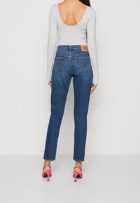 Jeans de mezclilla azul con un corte clásico, bolsillos traseros con detalles de costura y una camiseta de manga larga gris claro. Se pueden ver unos notables tacones rosas.