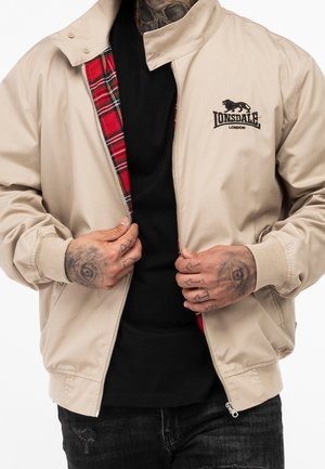 Hombre con chaqueta beige de Lonsdale London con forro de cuadros rojos, camiseta negra y manos tatuadas desabrochando parcialmente la chaqueta.