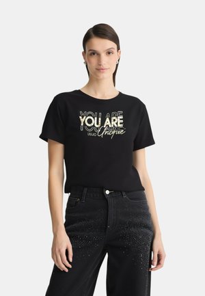 T-shirt in cotone nero con una stampa metallica dorata audace con il testo "SEI UNICO" in vari caratteri, abbinato a jeans in denim scuro con dettagli luccicanti.