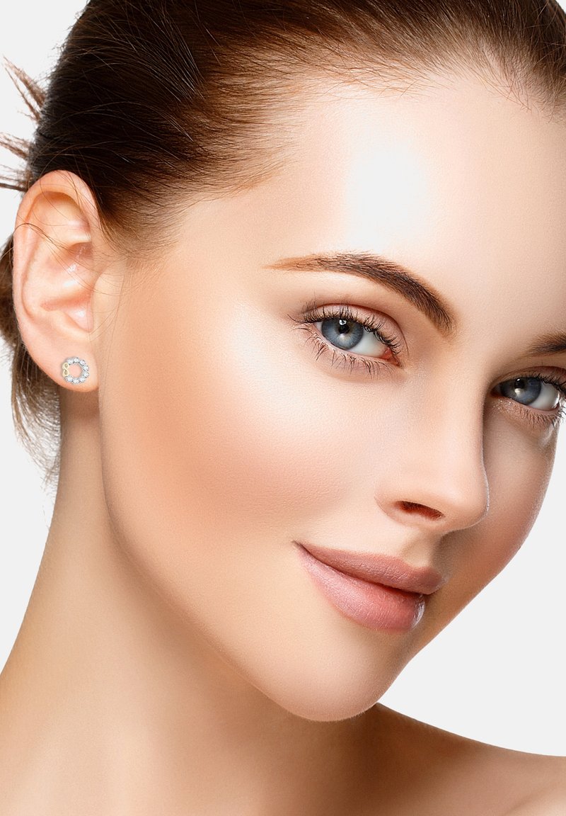 Närbild av en persons ansikte med slät, ljus hud och ljust brunt hår, prydd med runda studsörhängen med en klar sten i en silverföring.
