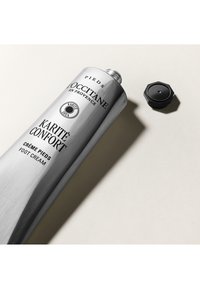 Aluminiowa tubka kremu do stóp L’Occitane, z wypukłym napisem, w kolorze szarym, z czarną sześciokątną nakrętką leżącą obok niej.