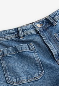 Mėlynos denim kelnių su mygtuko uždarymu, priekinėmis kišenėmis ir matomais siūlais; medžiaga turi tekstūruotą, šiek tiek išblukusią išvaizdą.