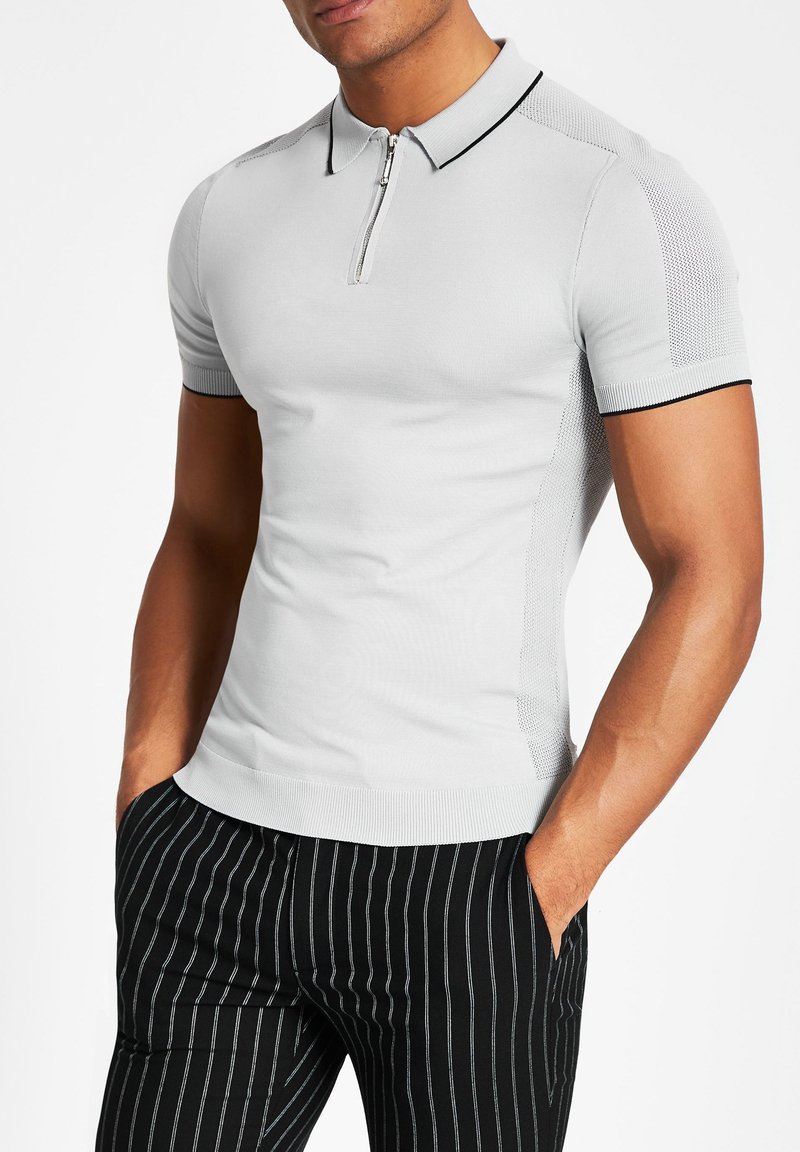 Homme portant un polo gris clair ajusté à manches courtes avec fermeture éclair à l'avant et un pantalon noir à rayures noires, mains dans les poches, sur fond blanc.