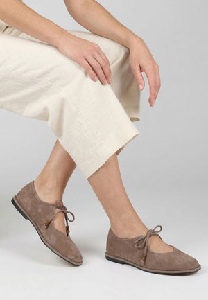 Person iført lyse beige kortbukser og taupe ruskind snørestøvler, siddende med hænderne hvilende på et knæ mod en ensfarvet baggrund.