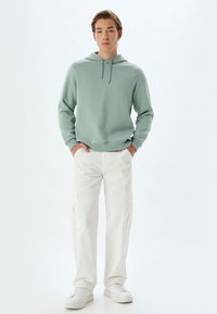 Sudadera verde claro con cordón, combinada con pantalones cargo blancos que cuentan con bolsillos. Calzado: zapatillas blancas. Diseño sencillo, look casual.