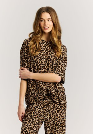 Jonge vrouw met lang golvend haar, gekleed in een bijpassende luipaardprint top en broek, staand met één arm over haar lichaam heen.