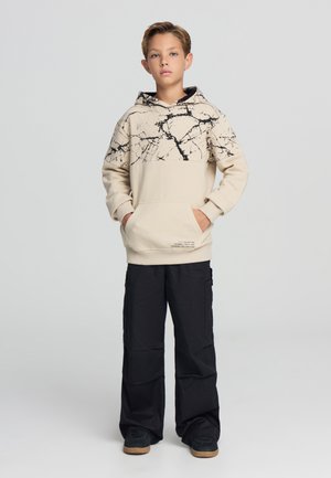 Hoodie beige con motivo in marmo nero sulla metà superiore, tasca a marsupio e orlo a costine; abbinato a pantaloni cargo neri e sneakers nere.
