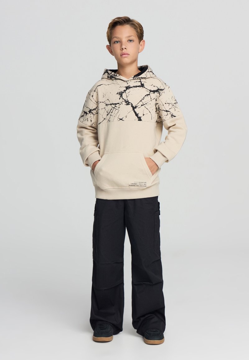 Hoodie beige con motivo in marmo nero sulla metà superiore, tasca a marsupio e orlo a costine; abbinato a pantaloni cargo neri e sneakers nere.