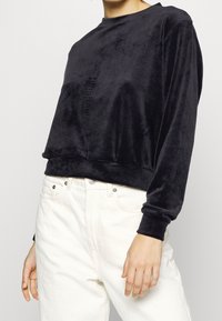 Svart, mjuk sammets sweatshirt med rund hals och långa ärmar; kortad nederkant; kombinerad med vita jeans. Subtil textur synlig på tyget.