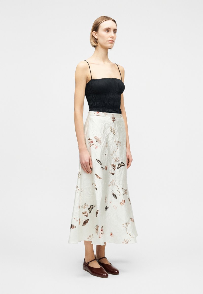 Woman wearing a black spaghetti strap top, white floral midi skirt, and dark brown Mary Jane flats, standing against a plain background.