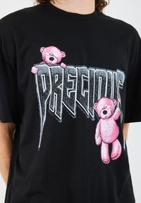 T-shirt di cotone nero con una grafica che rappresenta due orsi rosa che sorreggono la parola "PRECIOUS" in una scritta argentata stilizzata.