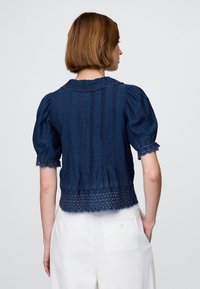 Mörkblå, texturerad blus med puffade ärmar, spetskant och vertikala veck. Har dekorativa utskurna mönster längs fållen.