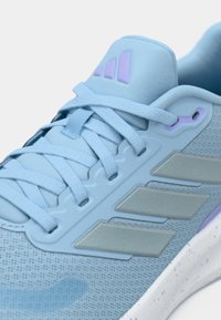 Svijetloplata sportska cipela s mrežastim gornjim dijelom, teksturiranim detaljima, toniranim vezicama i bijelim đonom s mrljama. Na boku ima tri sive pruge.