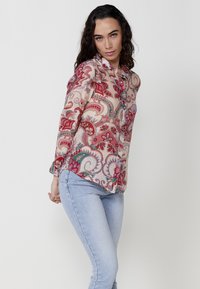 Blusa trasparente a maniche lunghe di un leggero beige con vivace motivo paisley in rosso, teal e rosa. Presenta una chiusura frontale con bottoni e un orlo arrotondato.