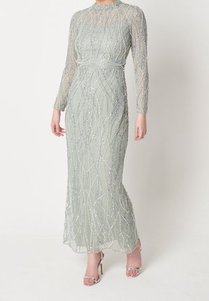 Pitkä hopeanvärinen mekko, jossa on läpikuultavat hihat ja monimutkaiset helmikuviot, yhdistettynä hopeisiin korkokenkiin. Malli seisoo yksiväristä taustaa vasten.