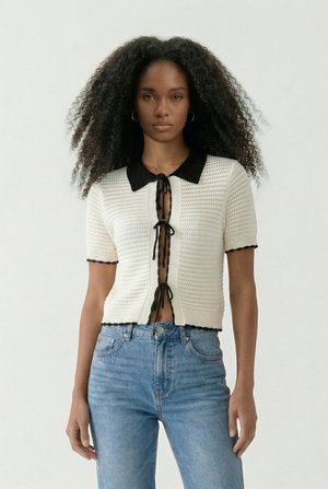 Femme aux longs cheveux bouclés portant un haut en maille blanc à manches courtes avec col noir et liens, associé à un jean bleu taille haute.