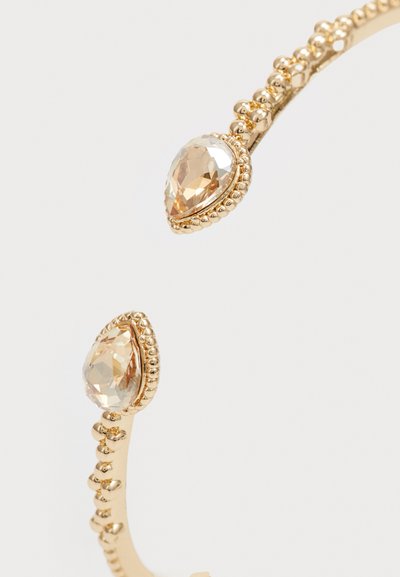 Pulsera de puño en tono dorado con dos cristales facetados en forma de pera rodeados por un diseño de cuentas texturizadas. Banda de metal lisa.