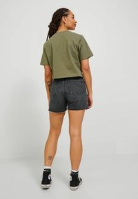 Camiseta verde oliva de corte ajustado, pantalones cortos de mezclilla negros con dobladillo desflecado y zapatillas negras de caña alta. Tatuaje floral visible en la parte inferior de la pierna.