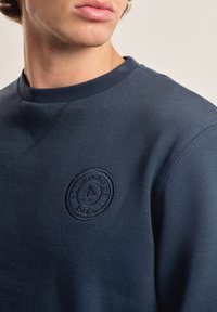 Sudadera azul marino con cuello redondo, que presenta un logotipo circular texturizado en color a juego en el pecho. Tejido suave y liso.