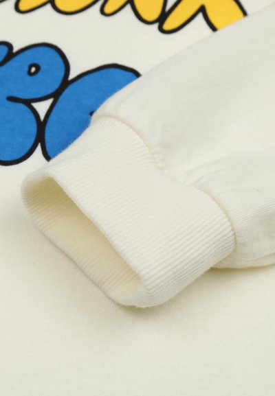 Gros plan sur un poignet de sweat-shirt crème à texture côtelée posé sur un tissu avec une impression graphique bleu et jaune audacieuse.