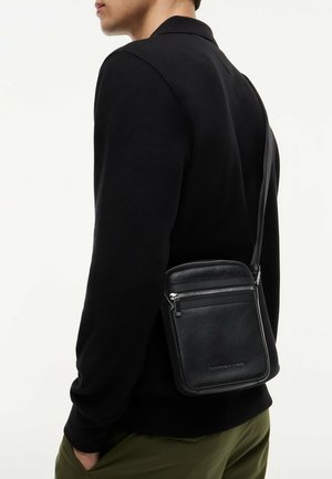 Mand iført sort sweater og olivengrønne bukser med en sort læder crossbody taske med lynlåse på rum.