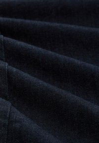 Tissu en denim bleu foncé avec une texture lisse, présentant des plis diagonaux et des nuances subtiles d'indigo. Coutures visibles le long des bords.