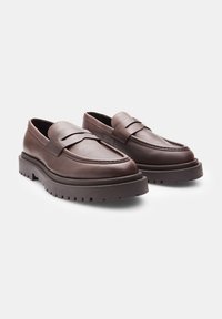 Mocassins en cuir marron avec une semelle plate, dotés d'un design cousu et d'une large bande sur le dessus. Semelle en caoutchouc texturée pour une meilleure adhérence.