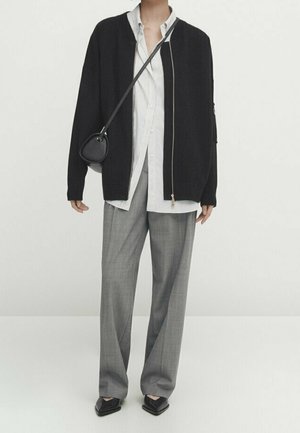 Personne portant une veste noire zippée sur une chemise blanche à rayures, un pantalon gris à jambes larges, des chaussures noires pointues et un petit sac bandoulière noir.