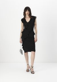 Black fitted dress with ruffle detailing along the front and shoulders, made of stretchy fabric. Worn with strappy sandals and a small metallic purse.