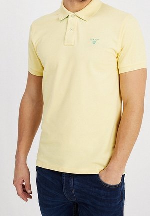 Mężczyzna w jasnopomarańczowej koszulce polo z krótkim rękawem i logo GANT na piersi, ubrany w ciemnoniebieskie dżinsy, z ręką w kieszeni.
