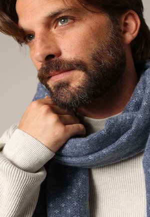 Écharpe texturée bleu marine enroulée autour du cou ; doux pull grisé tricoté avec des poignets côtelés. Gros plan sur un visage avec barbe et texture de peau subtile.